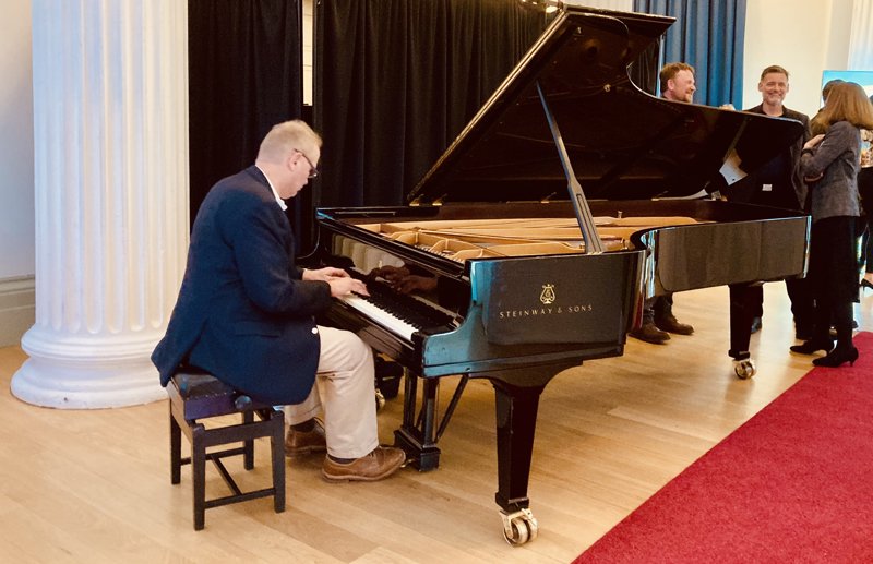 Man sitting on a stool playing a piano with three people listening